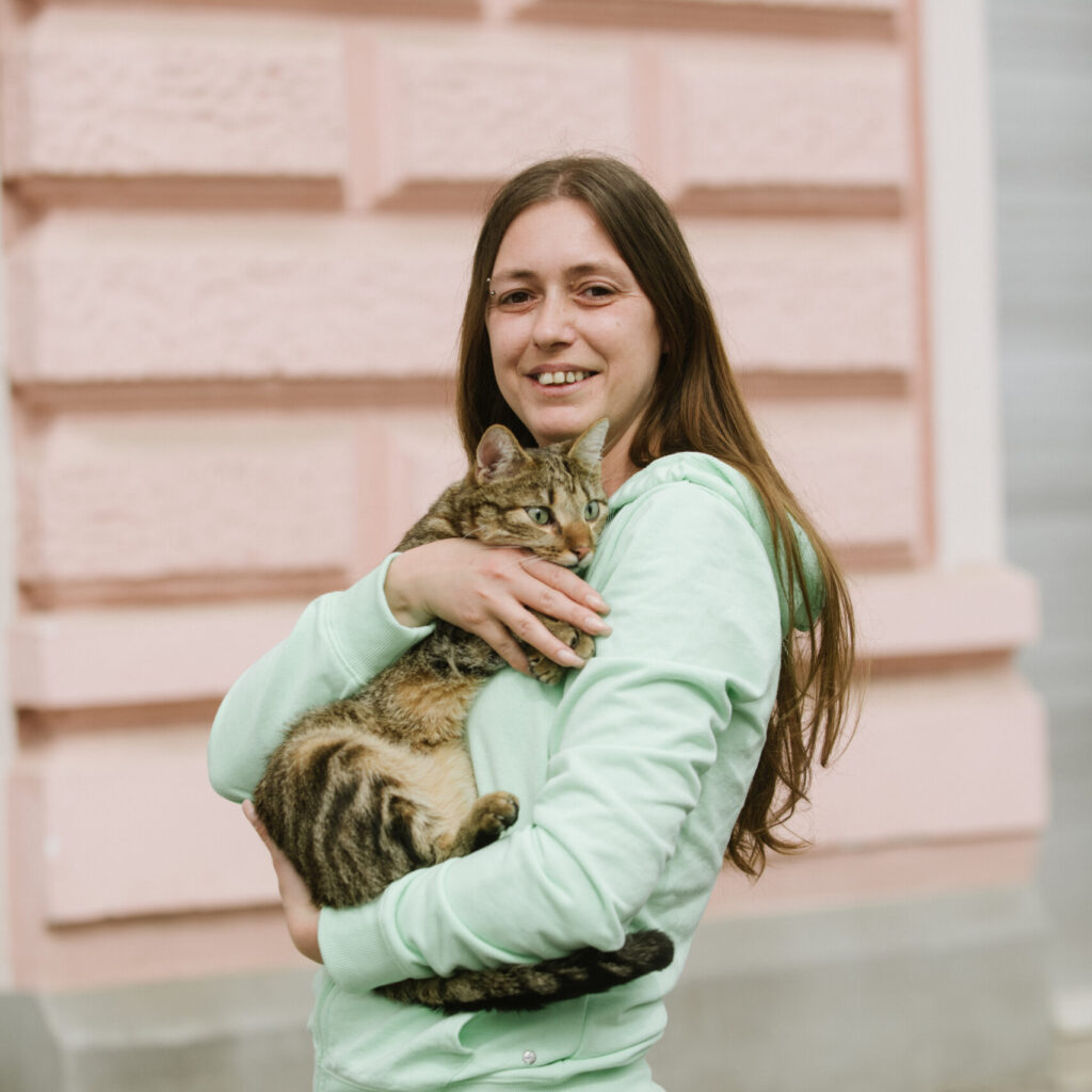 Lydia trägt einen grünen Pulli und hält ihre Katze im Arm. Sie steht draußen vor der Fassade eines Hauses.