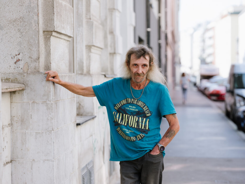 Josef P. ist Bewohner der neunerhaus Kudlichgasse. Im blauen Kurzarm-Tshirt lehnt er an einer Hausmauer, er trägt sein langes, graues Haar offen.