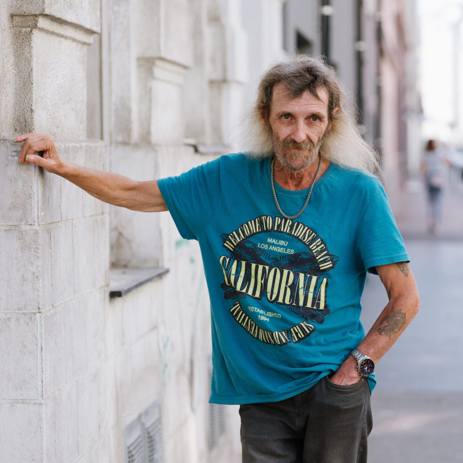 Josef P. ist Bewohner der neunerhaus Kudlichgasse. Im blauen Kurzarm-Tshirt lehnt er an einer Hausmauer, er trägt sein langes, graues Haar offen.